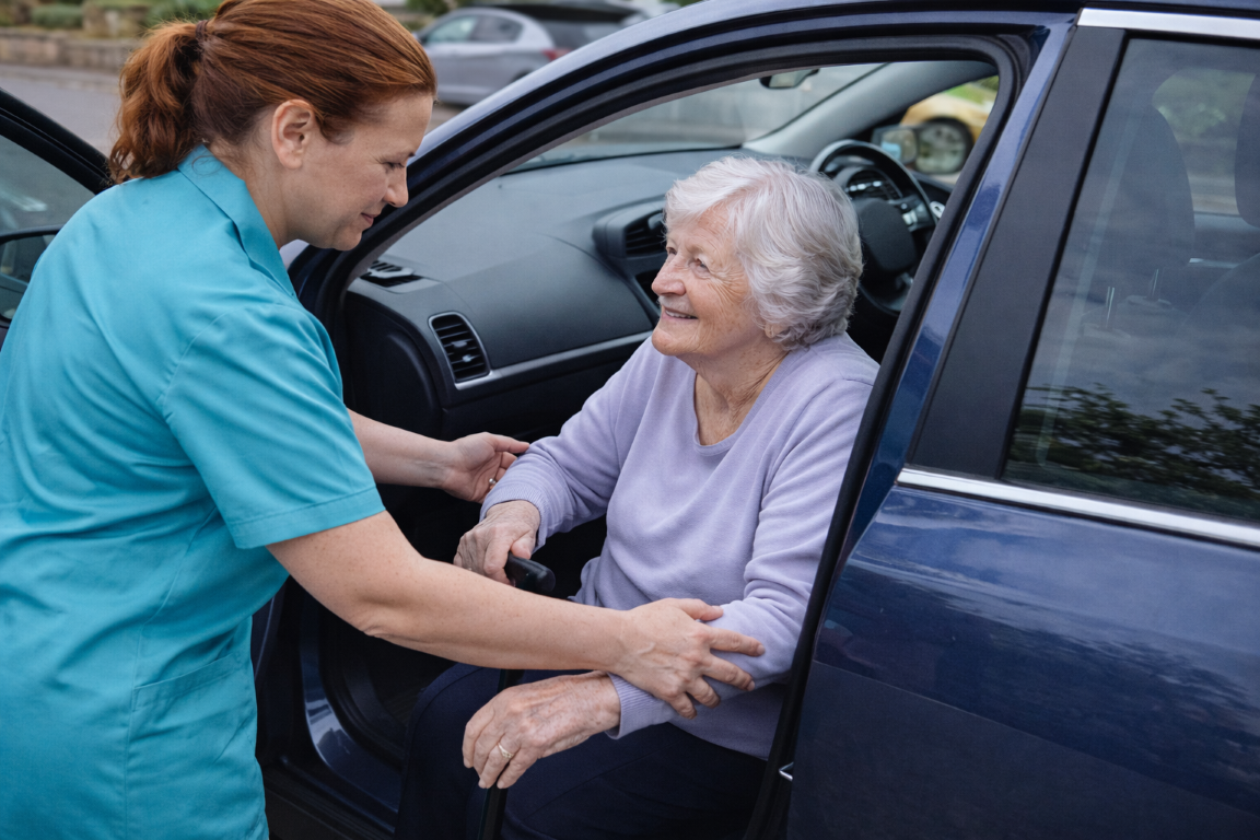 A carer in scrubs helps an elderly woman with grey hair out of a car, holding her arm supportively. The elderly woman is smiling and sitting in the front seat of the blue car. Helping her access an appointment.